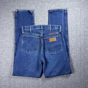 May include: Blue denim jeans with a classic five-pocket design. The jeans feature a straight leg cut and a brown leather patch on the back pocket with the Wrangler logo. The back pockets have a signature W-shaped stitching.