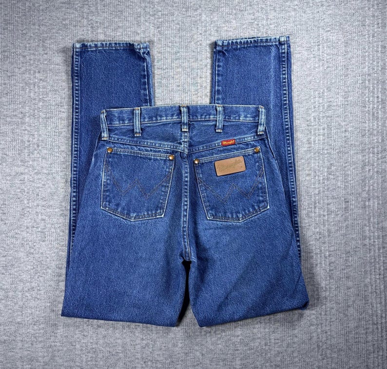 May include: Blue denim jeans with a classic five-pocket design. The jeans feature a straight leg cut and a brown leather patch on the back pocket with the Wrangler logo. The back pockets have a signature W-shaped stitching.