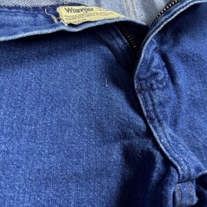 K&ouml;nnte beinhalten: Nahaufnahme einer blauen Jeans mit sichtbarem Rei&szlig;verschluss und dem Wrangler-Etikett. Die Jeans hat eine mittlere Waschung und weist Gebrauchsspuren auf. Das Etikett ist hellbraun mit schwarzem Text.