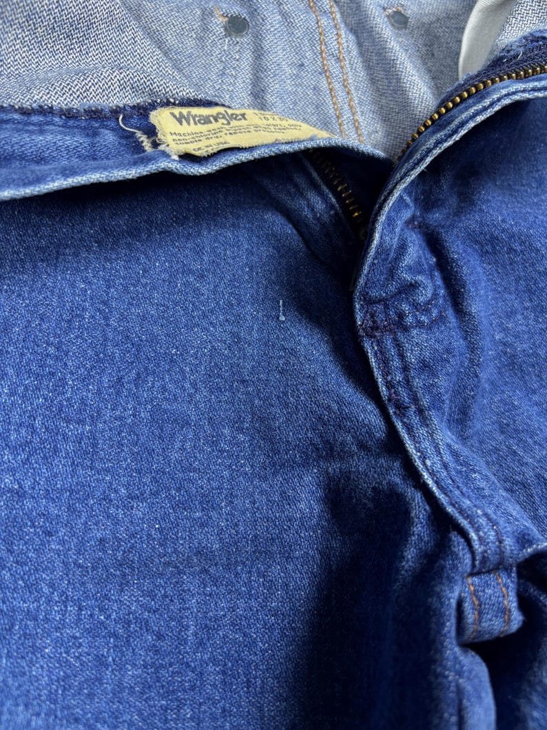 K&ouml;nnte beinhalten: Nahaufnahme einer blauen Jeans mit sichtbarem Rei&szlig;verschluss und dem Wrangler-Etikett. Die Jeans hat eine mittlere Waschung und weist Gebrauchsspuren auf. Das Etikett ist hellbraun mit schwarzem Text.