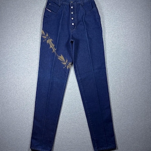 May include: Dark blue denim jeans with a high waist and button fly. The jeans feature a decorative gold-colored design on the left leg. The jeans have a classic five-pocket design.