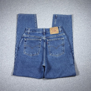 May include: A pair of blue denim jeans featuring a high-waisted design with two back pockets, a tan leather patch on the waistband, and a small red tab on the right pocket. The jeans have a classic straight-leg cut with visible stitching.