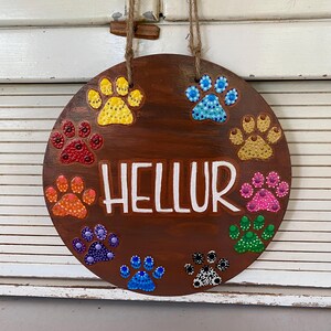 May include: A round, wooden sign with a dark brown stain, featuring colorful paw prints in yellow, red, orange, purple, blue, pink, green, and gold. The word "HELLUR" is painted in white across the center. The sign hangs by a jute rope.