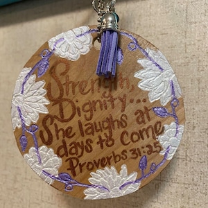 May include: A round wooden ornament with a purple tassel and white flowers painted around the edge. The ornament has the text "Strength, Dignity... She laughs at days to come Proverbs 31:25" written on it.