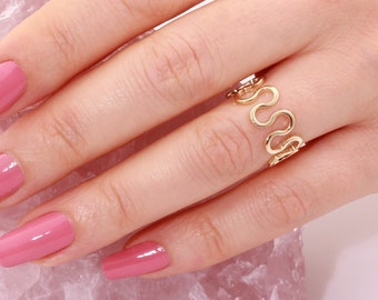 Pode incluir: Um anel de ouro com um design ondulado abstrato. O anel está em uma mão com esmalte de unha rosa.