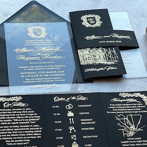 May include: Black and gold wedding invitation suite with a clear acrylic cover. The invitation features a crest with the couple's names and wedding date. The RSVP card includes a map of the wedding venue and a list of events for the day.