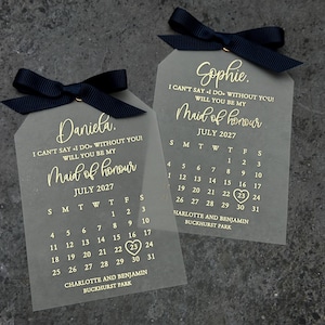 May include: Two clear acrylic tags with black ribbon bows. Each tag has gold lettering that says "I can't say I do without you! Will you be my Maid of honor" and a calendar for July 2027. The top tag says "Sophie." and the bottom tag says "Daniela."