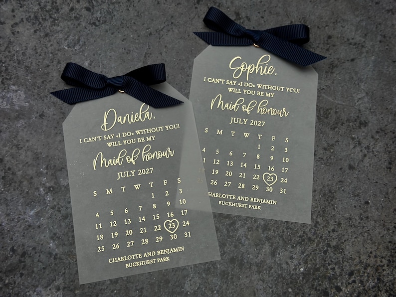 May include: Two clear acrylic tags with black ribbon bows. Each tag has gold lettering that says "I can't say I do without you! Will you be my Maid of honor" and a calendar for July 2027. The top tag says "Sophie." and the bottom tag says "Daniela."