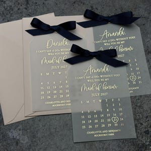 May include: Two clear acrylic tags with black ribbon bows. The tags feature a gold calendar with the month of July 2027 and the text "I can't say I do without you! Will you be my Maid of Honor."  The names "Amanda" and "Daniela" are printed on the tags.
