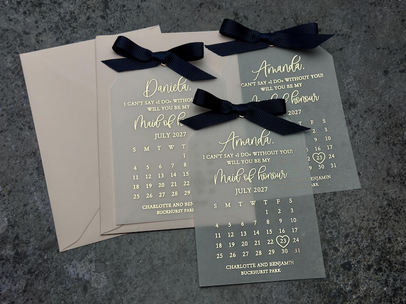 May include: Two clear acrylic tags with black ribbon bows. The tags feature a gold calendar with the month of July 2027 and the text "I can't say I do without you! Will you be my Maid of Honor."  The names "Amanda" and "Daniela" are printed on the tags.