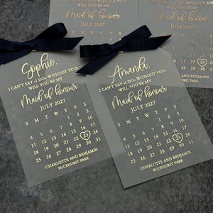 May include: Two clear acrylic tags with black ribbon and gold lettering. The tags feature a calendar for July 2027 with the text "Maid of honor" and the names "Sophie" and "Amanda".