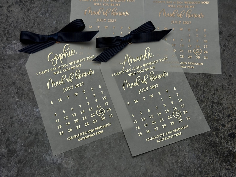 May include: Two clear acrylic tags with black ribbon and gold lettering. The tags feature a calendar for July 2027 with the text "Maid of honor" and the names "Sophie" and "Amanda".
