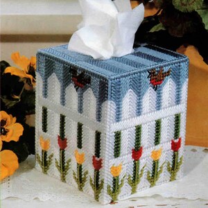 May include: A square tissue box cover, hand-stitched with a blue top and a white picket fence design. The sides are decorated with red and yellow tulips. Two small red birds are on the top. White tissues are visible.