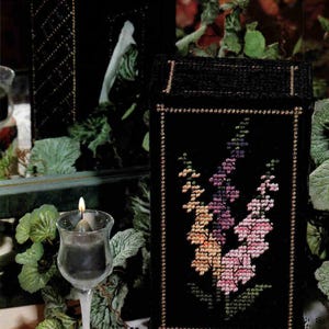 May include: Black tissue box cover and matching decorative box with gold trim. A black needlepoint piece features a floral design in shades of pink, purple, and yellow. A lit candle in a glass holder sits nearby, adding a warm glow.