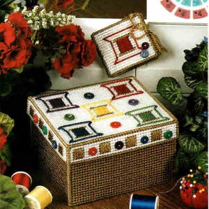 May include: A handcrafted, square storage box with a lid, featuring a white background with colorful square designs and buttons. The box has a brown woven base and is accompanied by a smaller matching square item. Sewing notions are visible.