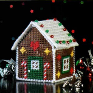 May include: A handcrafted Christmas decoration in the shape of a gingerbread house. The house is brown with a white roof, decorated with red, green, and white accents. Candy canes and a heart adorn the front. The house is approximately 4 inches tall.