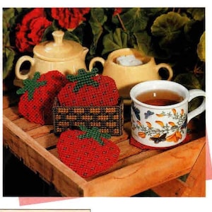 May include: A still life arrangement featuring a cup of tea, strawberry-shaped coasters, and a sugar bowl. The coasters are red with green tops, and the cup is white with a floral design. The sugar bowl is yellow.