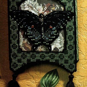 Puede incluir: Un tapiz de pared decorativo hecho a mano con un diseño de mariposa negra con detalles de cuentas. La mariposa está colocada sobre un fondo de encaje floral dentro de un marco verde bordado. Cuelgan borlas negras en la parte inferior.