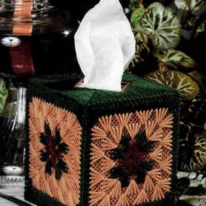 May include: A handmade tissue box cover in a square shape. The box is dark green with a tan and burgundy geometric design on each side. White tissues are visible at the top. The box is sitting on a patterned surface.