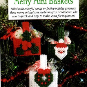 May include: Three small plastic canvas baskets with handles, each containing wrapped candies. One red basket features a green holly motif, one dark green basket displays a Santa face, and one white basket shows a green wreath with a red bow.