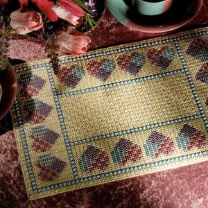 May include: A rectangular placemat featuring a woven tan base with a grid pattern. The border is decorated with a repeating motif of small, multi-colored hearts in shades of red, purple, and blue, arranged in a structured, geometric layout.