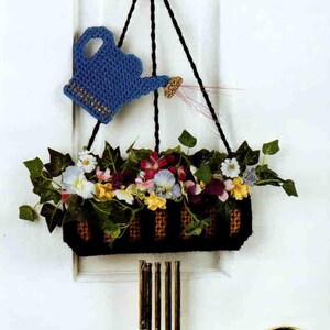 May include: A crocheted blue watering can wall hanging with a black basket containing artificial flowers and green leaves. Five metallic gold wind chimes hang from the bottom of the basket, suspended by black yarn cords against a white door.