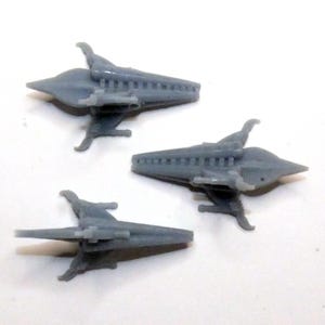 May include: Three small, gray spaceship models are arranged on a white surface. Each model has a pointed front, wings, and a detailed body, suggesting a futuristic or science fiction theme. The models are likely for display or a hobby.
