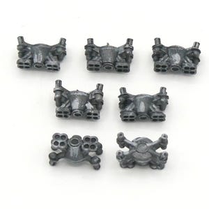 May include: Seven small, metallic gray model parts are arranged on a white background. The parts have a complex, industrial design with multiple nozzles and a central body. The objects appear to be components of a larger model or machine.