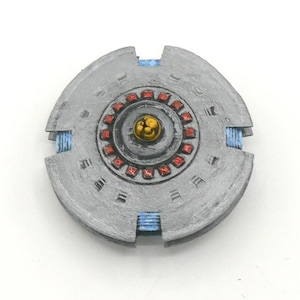 May include: A gray, circular object with a central, red-accented design and a gold-colored center. The object has blue accents and a textured surface, resembling a futuristic or mechanical component.