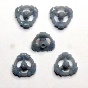 May include: Five small, gray, plastic wheel-shaped components. Each component has a central hub with three spokes extending to a circular outer rim. The rims have a series of small, evenly spaced protrusions.