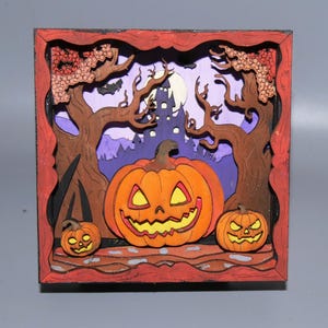 May include: A wooden Halloween-themed shadow box featuring a carved pumpkin with a smiling face, flanked by two smaller jack-o'-lanterns. The background includes a spooky castle, trees, and a full moon, all within a red frame.