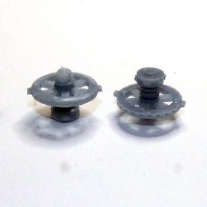 May include: Two small, gray plastic components. Each has a circular, slotted top with a central post and a base. The components are on a white surface, casting soft shadows.