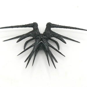 May include: A dark, textured sculpture resembling stylized wings, with multiple sharp, pointed projections. The wings are symmetrical and appear to be made of a dark, possibly metallic material, set against a white background.