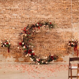 May include: A brick wall with a floral archway made of red, pink, and white flowers. The archway is decorated with greenery and vines. There are two small floral arrangements on either side of the archway. The floor is covered in red rose petals.