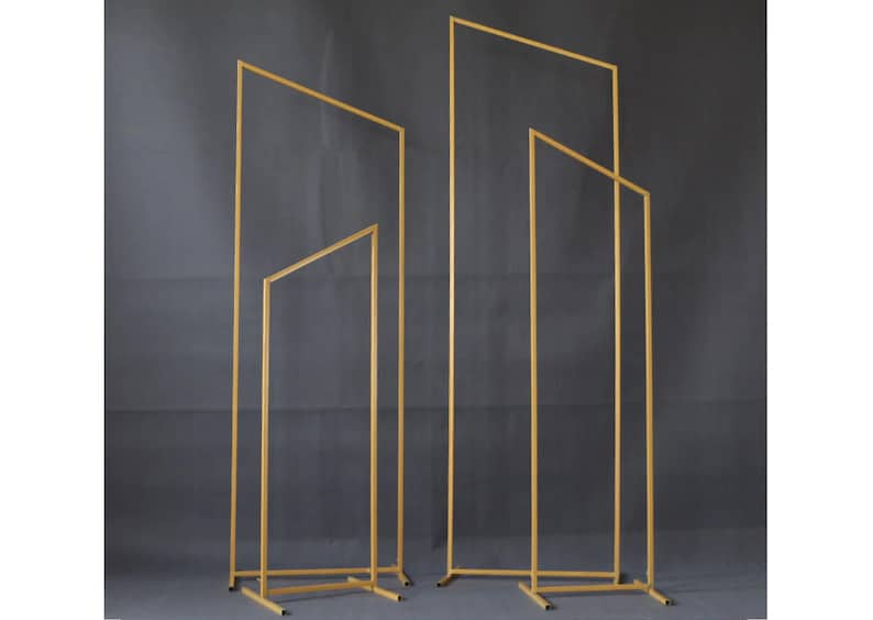 May include: Four gold metal geometric arch stands of varying heights are displayed against a dark grey backdrop. The stands have a rectangular base and a linear design, suitable for weddings or events.