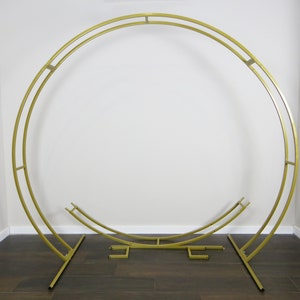 May include: A gold metal archway with a circular design. The archway is made of two separate pieces that can be assembled together. The archway is standing on a wooden floor.