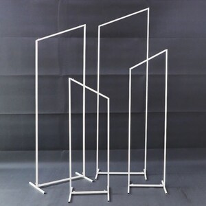 May include: Four white metal rectangular frames of varying heights, set against a dark grey backdrop. The frames are made from thin, straight bars, creating a minimalist, geometric design. The frames stand upright, supported by rectangular bases.