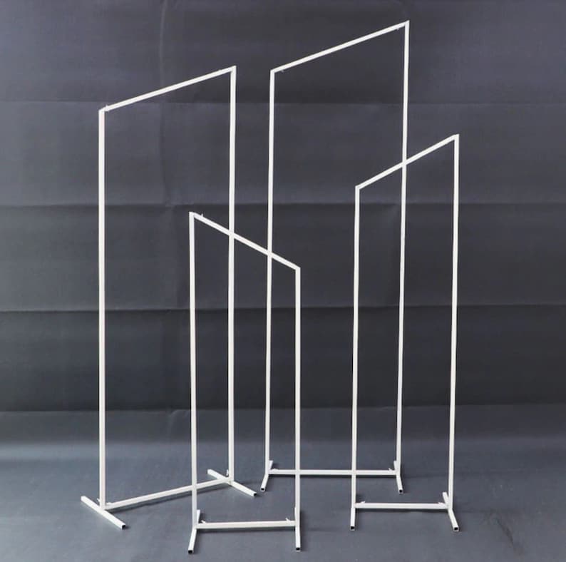 May include: Four white metal rectangular frames of varying heights, set against a dark grey backdrop. The frames are made from thin, straight bars, creating a minimalist, geometric design. The frames stand upright, supported by rectangular bases.