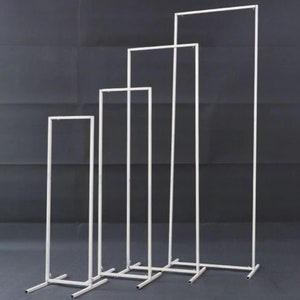 May include: Four white metal display stands of varying heights are arranged in a row. Each stand has a rectangular frame and a base for stability. The stands are designed for displaying items.