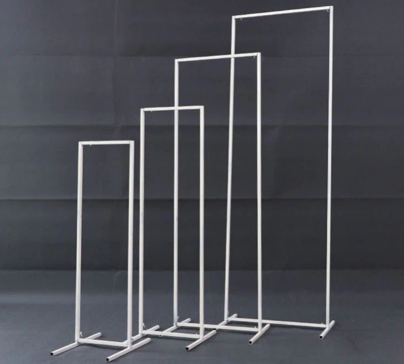 May include: Four white metal display stands of varying heights are arranged in a row. Each stand has a rectangular frame and a base for stability. The stands are designed for displaying items.