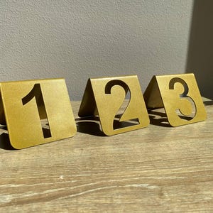 Puede incluir: Tres portanúmeros de mesa de metal dorado, cada uno con un número diferente: 1, 2 y 3. Los números están recortados del metal, creando un espacio negativo. Los soportes son triangulares y se apoyan sobre una superficie de madera.