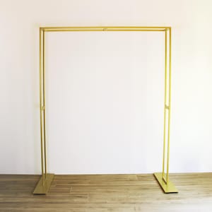 May include: A gold metal archway stand with a simple rectangular design. The stand is freestanding and has a flat base.