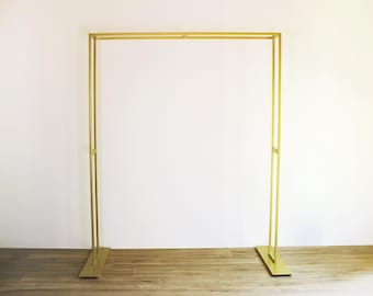 May include: A gold metal archway stand with a simple rectangular design. The stand is freestanding and has a flat base.