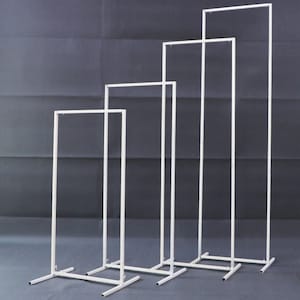 May include: Four white metal rectangular display stands of varying heights. Each stand has a rectangular base and a top bar, creating a frame. The stands are arranged in a row, with the tallest one on the right.