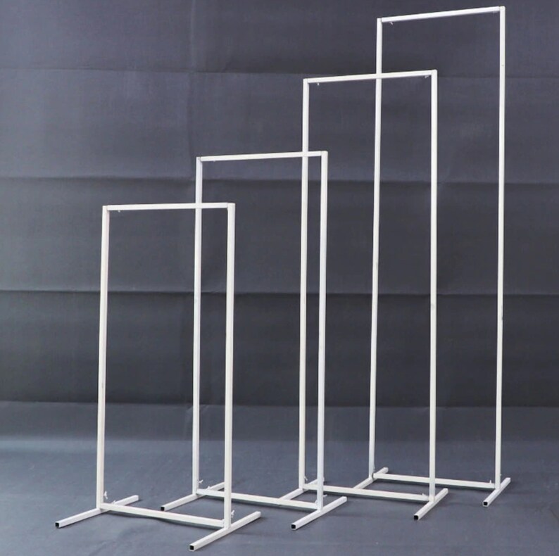 May include: Four white metal rectangular display stands of varying heights. Each stand has a rectangular base and a top bar, creating a frame. The stands are arranged in a row, with the tallest one on the right.