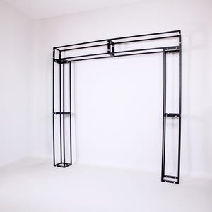 May include: A black metal archway structure with a rectangular top and vertical supports. The frame is made of thin, straight metal bars, creating a minimalist design. The structure is freestanding and set against a white background.