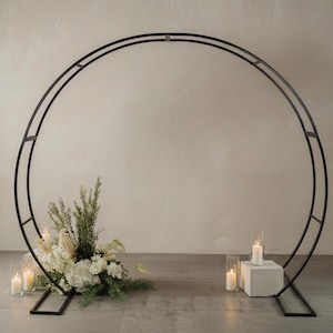 May include: A large, circular black metal arch frame with a double-ring design. The structure is supported by two rectangular base stands. A floral arrangement with white flowers and greenery sits at the base, accompanied by several lit pillar candles.