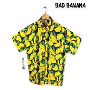 Pu&ograve; includere: Una camicia a maniche corte con bottoni con una stampa di banane gialle su uno sfondo blu scuro. La camicia ha una tasca sul lato sinistro e il testo "BAD BANANA" &egrave; stampato nell'angolo in alto a destra.