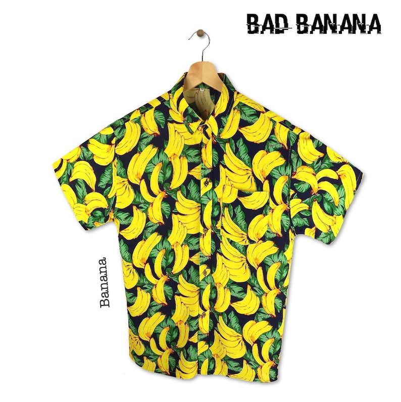 Pu&ograve; includere: Una camicia a maniche corte con bottoni con una stampa di banane gialle su uno sfondo blu scuro. La camicia ha una tasca sul lato sinistro e il testo "BAD BANANA" &egrave; stampato nell'angolo in alto a destra.