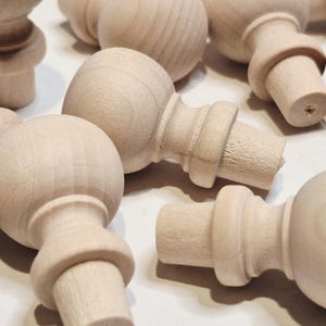 May include: A collection of unfinished wooden furniture finials. Each finial features a rounded top, a decorative midsection, and a cylindrical base. The natural wood grain is visible on the surface of the finials.